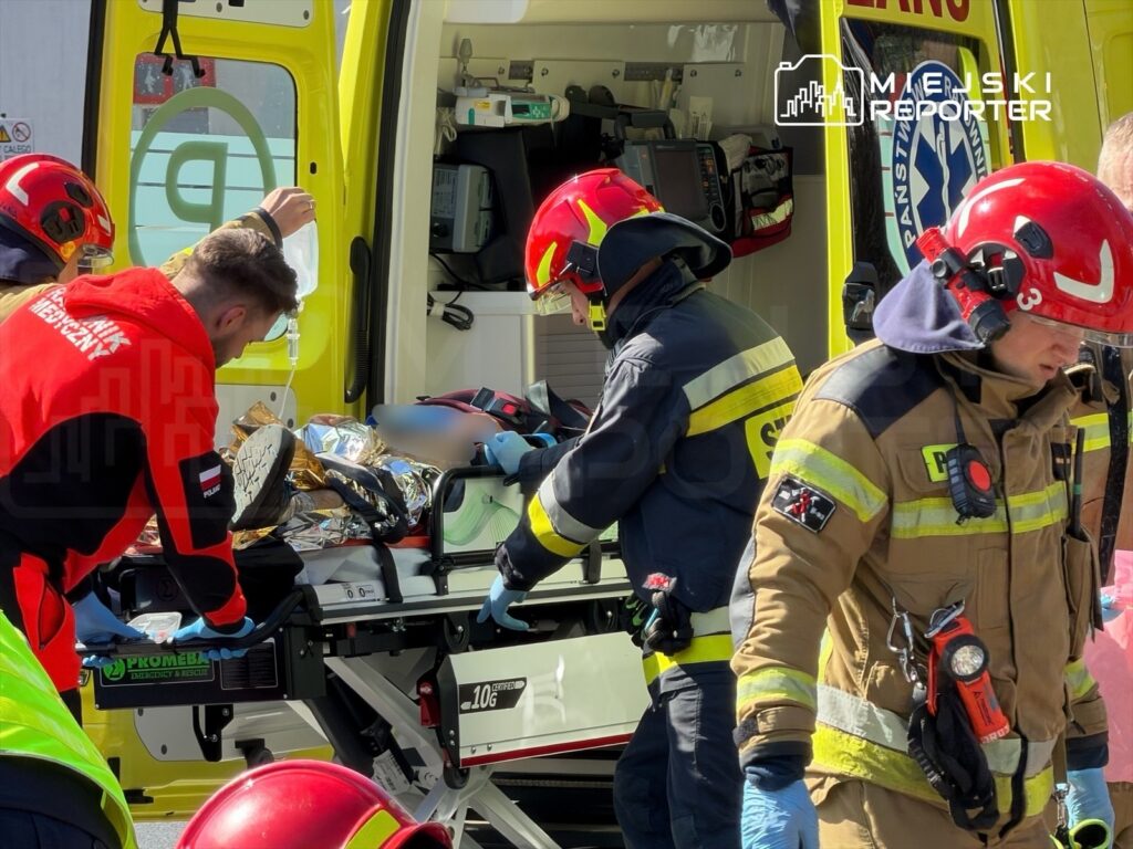 Ratownicy medyczni transportują poszkodowanego na noszach do karetki, pracując w słonecznym otoczeniu.
