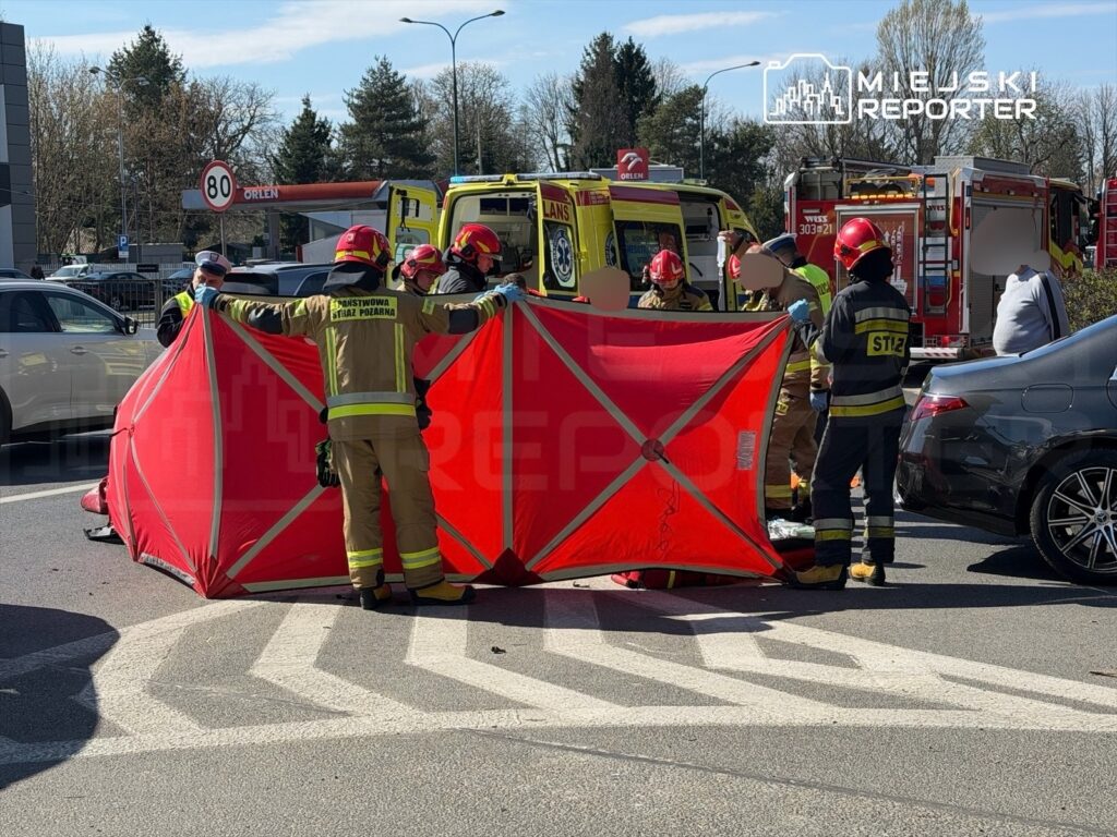 Grupa strażaków ustawia czerwoną osłonę na skrzyżowaniu, podczas akcji ratunkowej po wypadku drogowym.