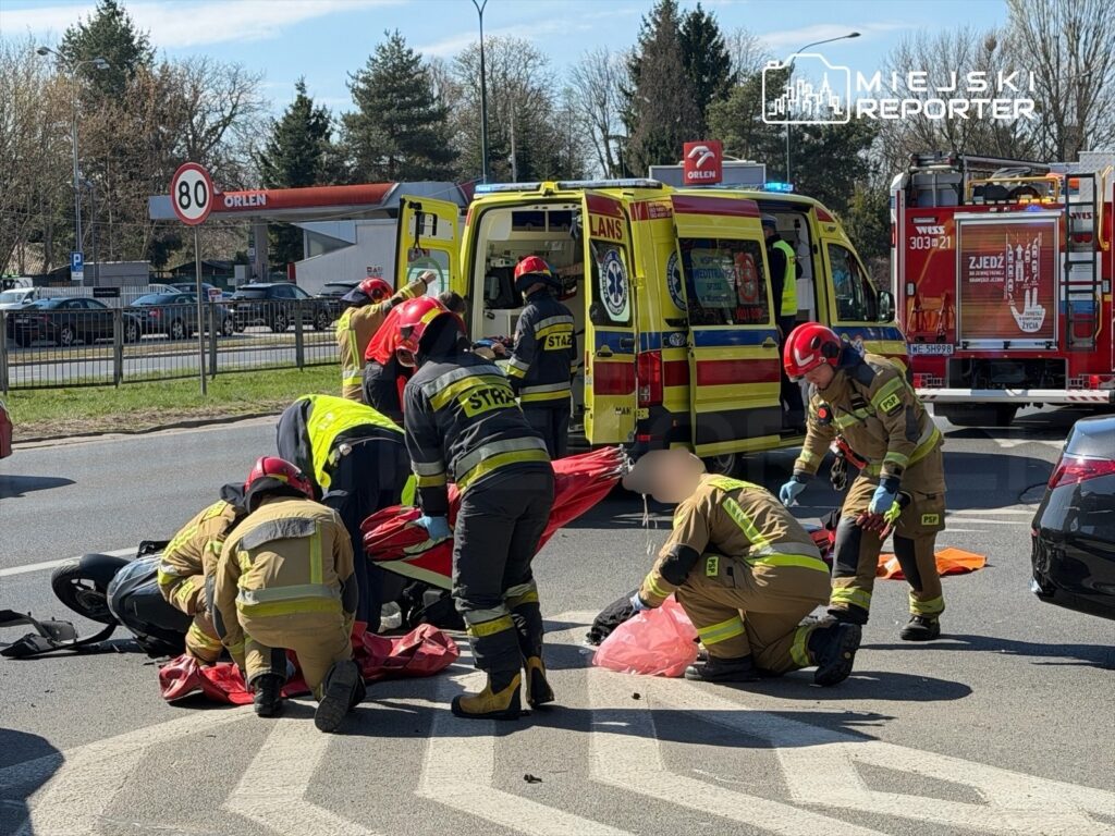 Grupa strażaków w żółtych i czarnych mundurach udziela pomocy poszkodowanemu na drodze, obok stoją karetki i wóz strażacki.