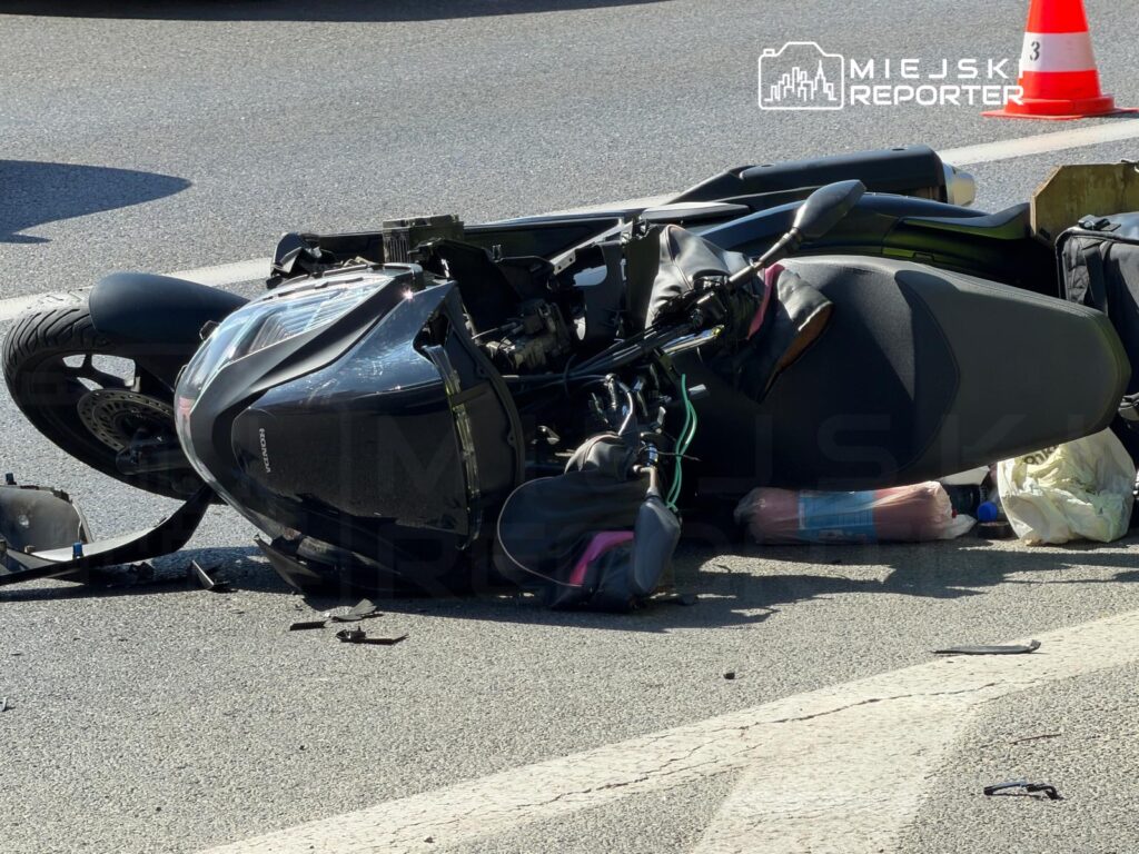 Przewrócony motocykl leży na jezdni, obok widoczne fragmenty pojazdu oraz przedmioty osobiste.
