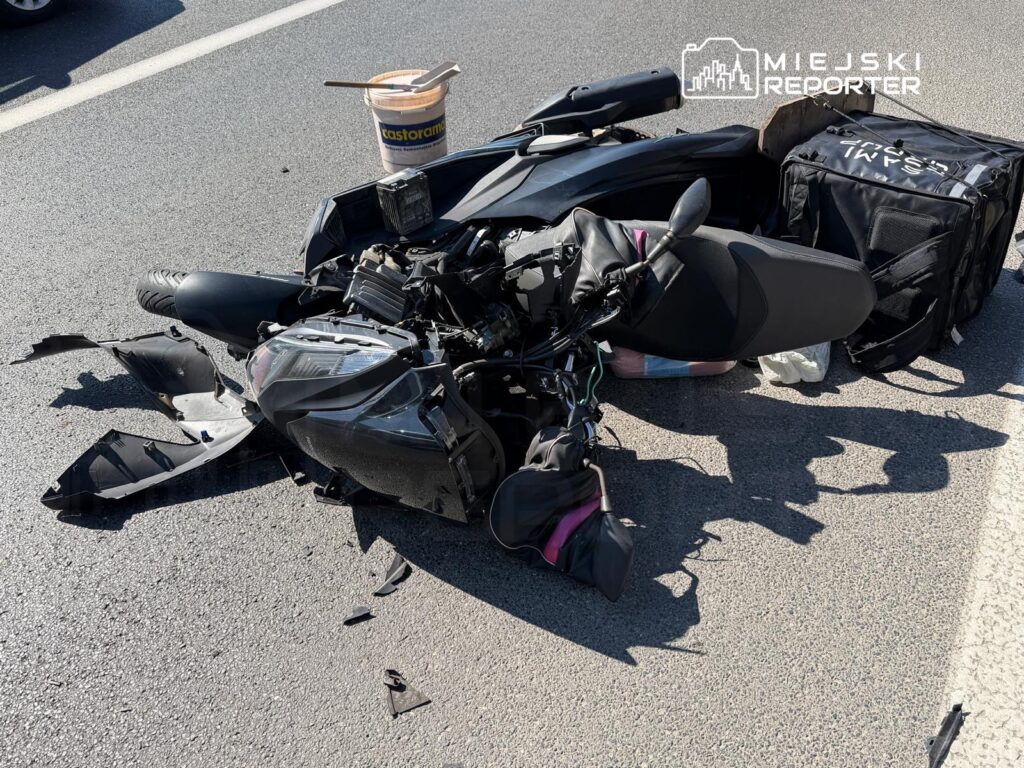 Motocykl leży na jezdni, z uszkodzonym przodem i rozrzuconymi elementami, obok znajduje się pojemnik z farbą.