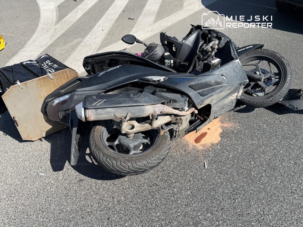 Przewrócony skuter leżący na asfalcie z rozlaną cieczą, otoczony znakami drogowymi.