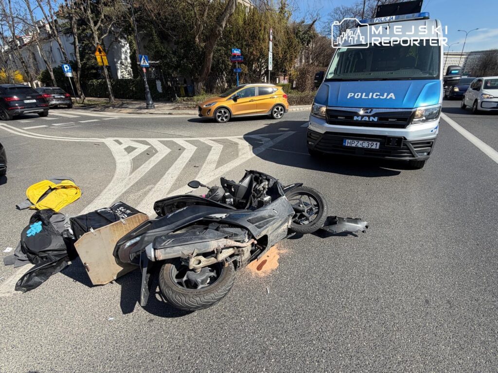 Zniszczony skuter leży na drodze obok policyjnego samochodu, a na jezdni leżą porzucone ubrania i torba.