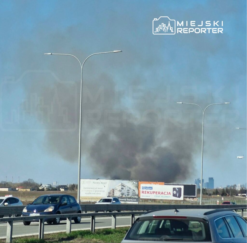 Czarny dym unoszący się nad horyzontem w tle ruchliwej drogi, obok billboardy reklamowe.