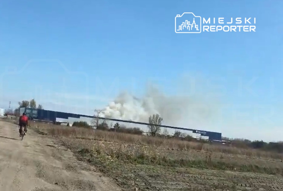 Rowerzysta jedzie po drodze, w tle widoczny dym unoszący się z przemysłowego budynku pod jasnym niebem.