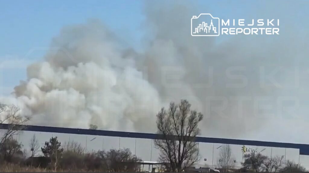 Gęsty, szary dym unoszący się nad nowoczesnym budynkiem z przeszkloną fasadą w słoneczny dzień.