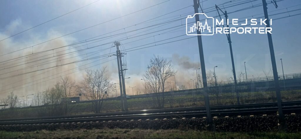 Gęsty dym unoszący się nad terenami przemysłowymi w pobliżu torów kolejowych w słoneczny dzień.