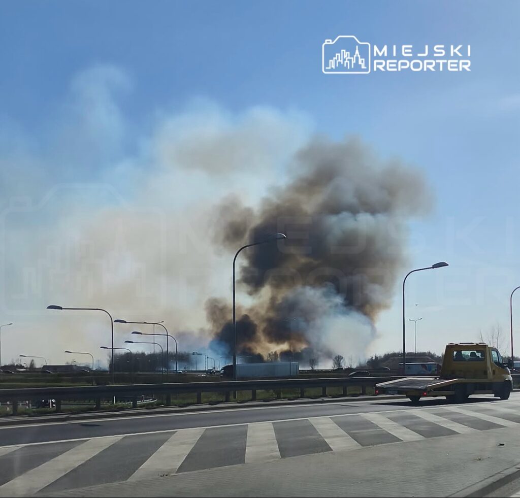 Gęsty dym unoszący się nad terenem przemysłowym w tle, widoczny z ruchliwej drogi.