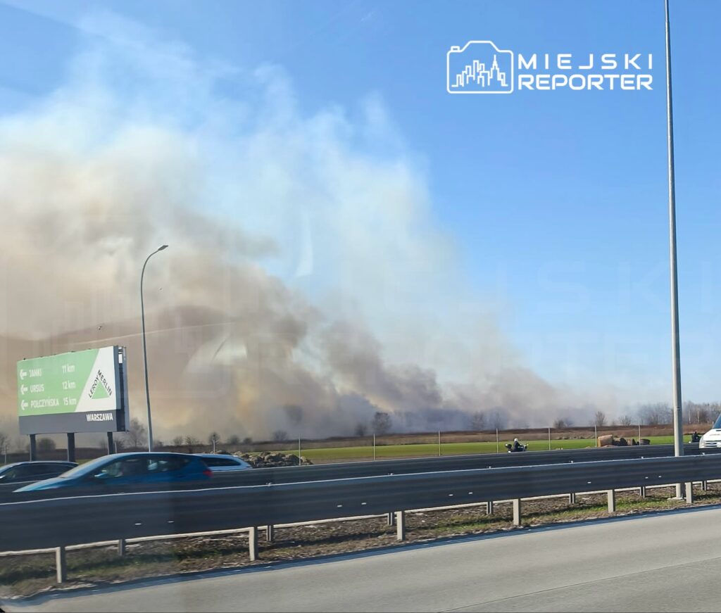 Gęsty dym unoszący się nad polem, widoczny z ruchliwej drogi, obok znak drogowy wskazujący kierunki.