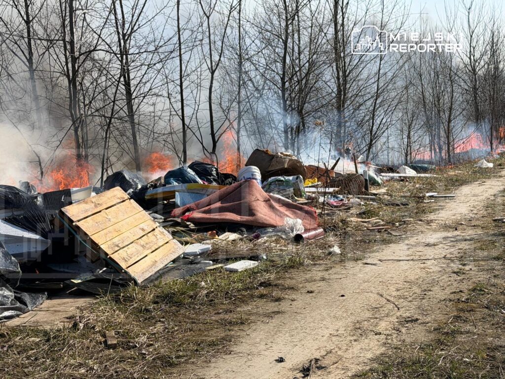 Ognisko płonące wśród sterty odpadów, w tle widoczne nagie drzewa i dym unoszący się w powietrzu.