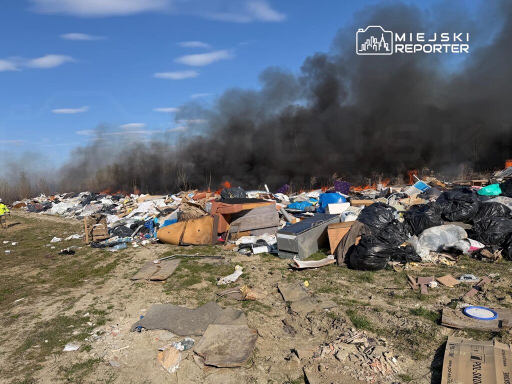 Wielka sterta odpadów płonąca na otwartym terenie, z unoszącym się czarnym dymem w tle.