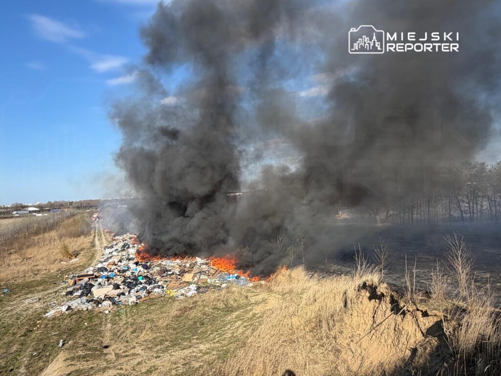 Ogień i gęsty czarny dym unoszą się nad stertą odpadów na otwartej przestrzeni obok ścieżki wśród traw.