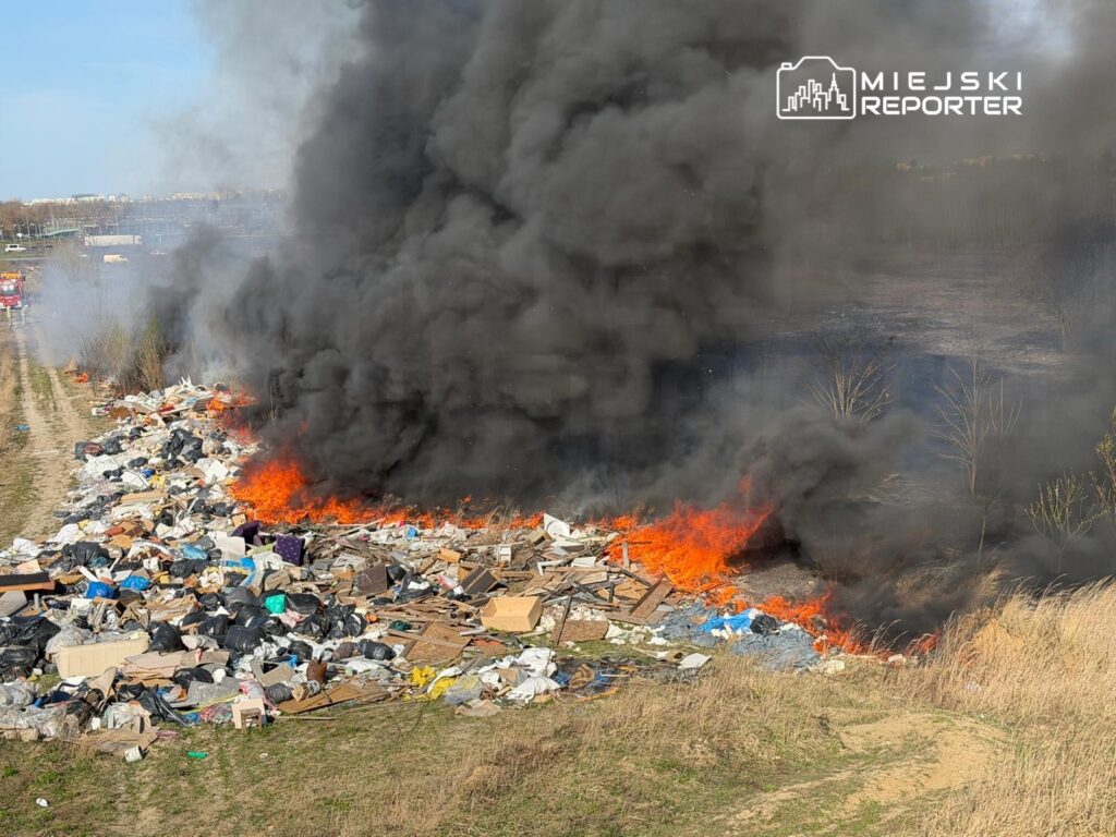 Ogień trawi stertę odpadów, wydobywając gęsty czarny dym, w tle widoczne pojazdy straży pożarnej.