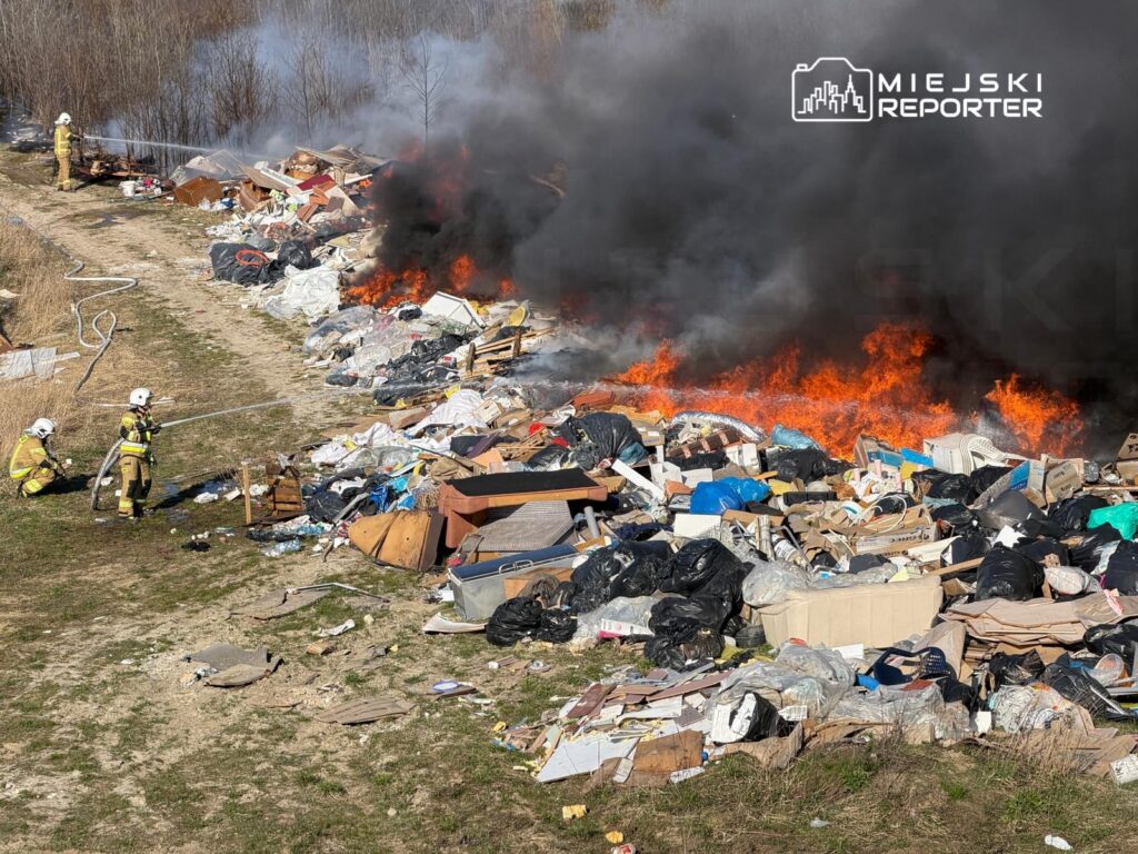 Strażacy gaszą pożar w hałdzie odpadów, wokół której unosi się gęsty dym i płomienie.