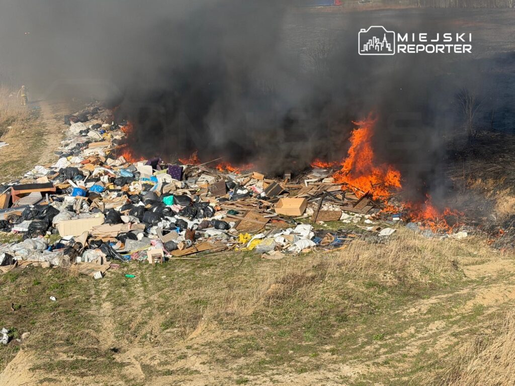 Ognisko z płonącymi odpadami wśród zarośli, obok drogi. W tle widoczny dym i fragmenty mebli.