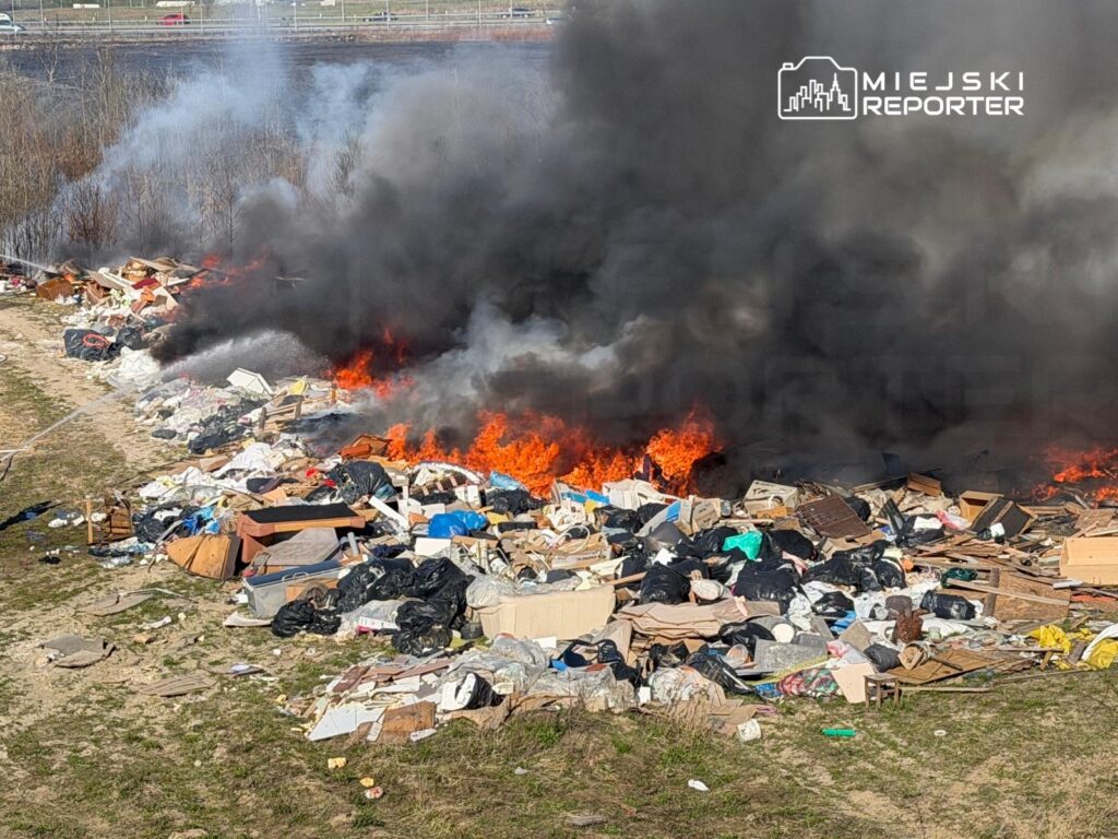 Ogień trawi stos odpadków na skraju pola, wydobywając gęsty dym i intensywne płomienie.