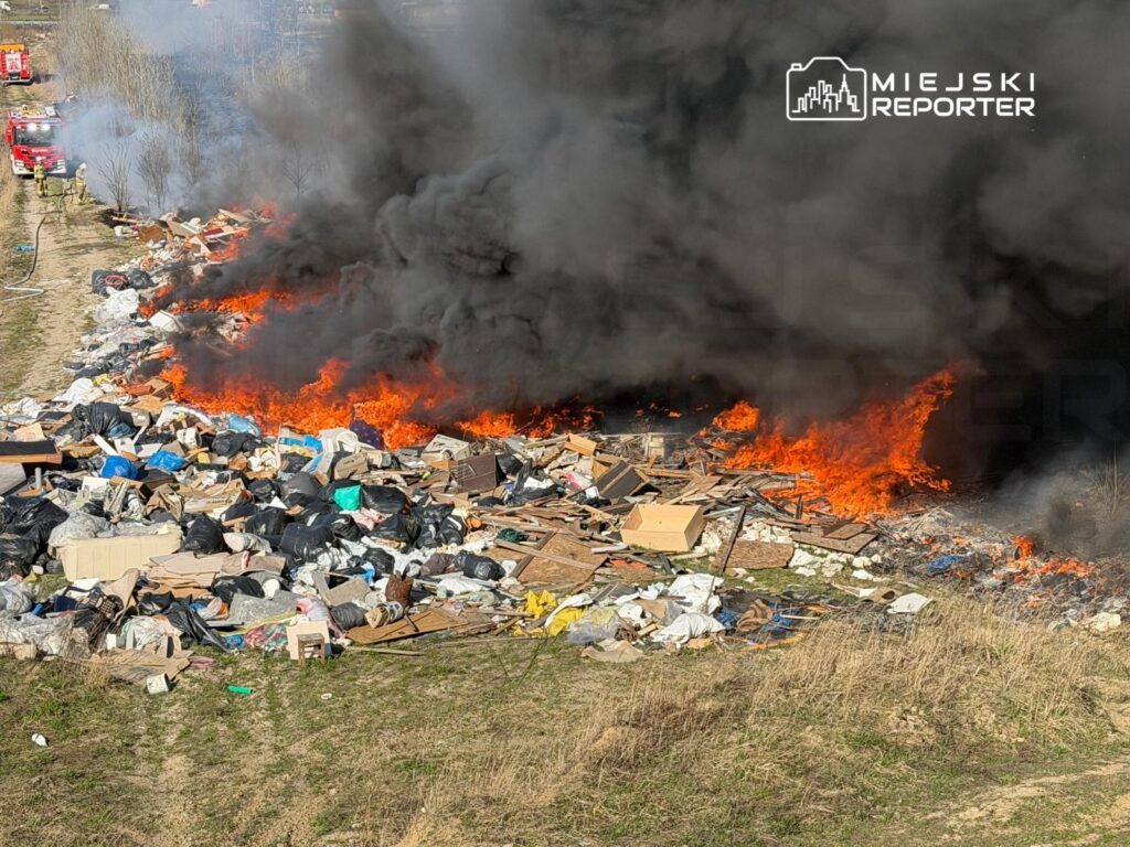 Wielka sterta odpadów płonąca na wysypisku, otoczona gęstym czarnym dymem, w tle widać wozy strażackie.