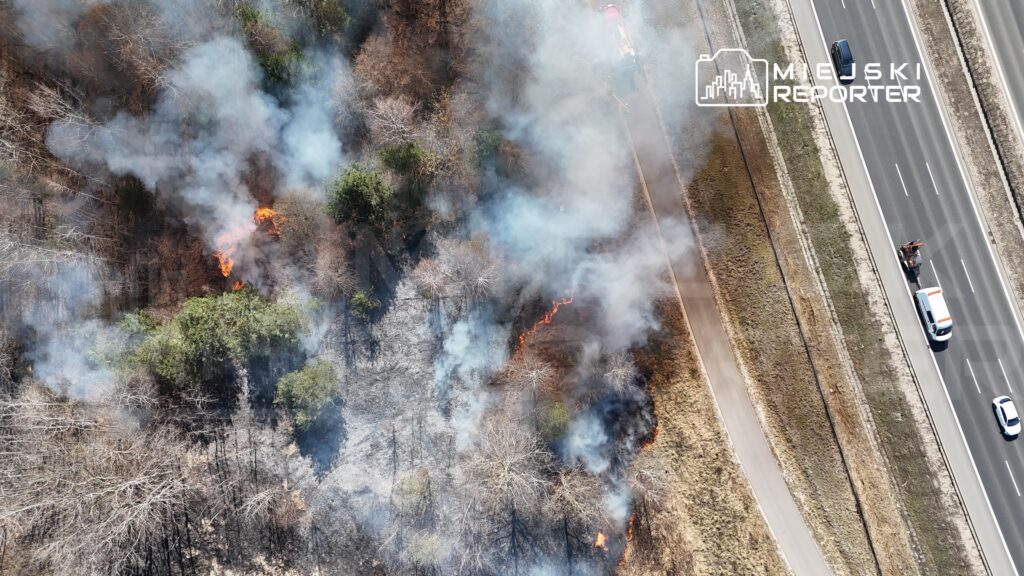 Pożar trawy i krzewów z płomieniami oraz dymem, blisko drogi, obok widoczne pojazdy służb ratunkowych.