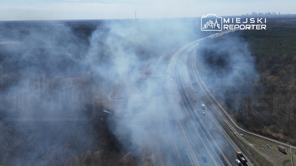 Dym unoszący się nad drogą, w tle widoczna zalesiona przestrzeń oraz fragmenty zabudowy.