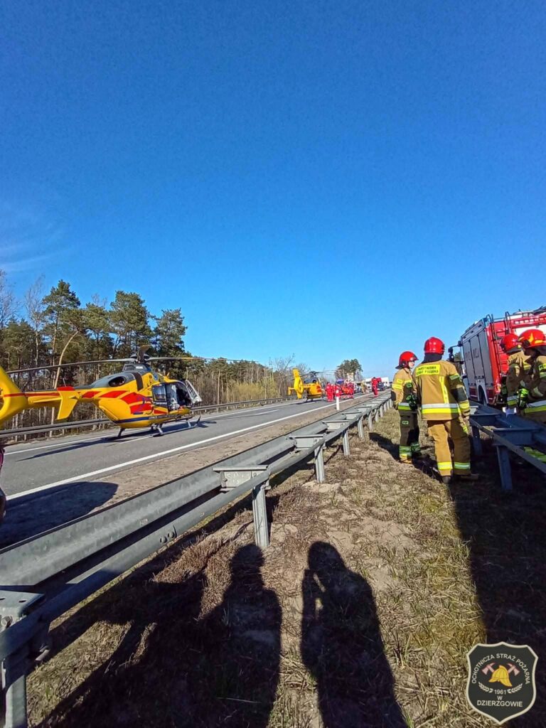 Grupa strażaków w mundurach pracuje przy drodze, obok lądowiska dla helikopterów ratunkowych.
