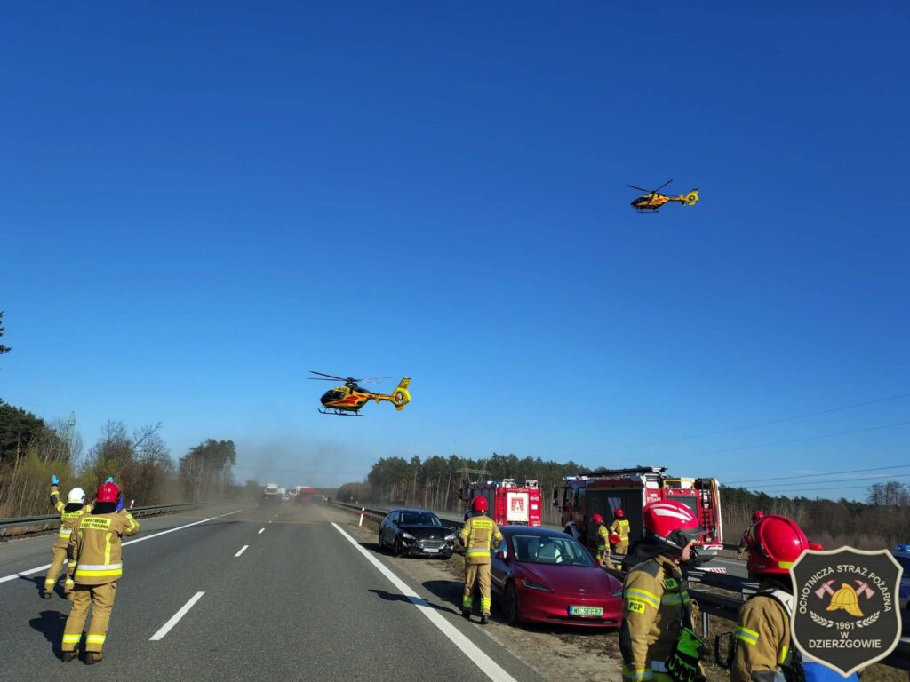 Dwóch strażaków w mundurach wskazuje kierunek, podczas gdy dwa śmigłowce ratunkowe unoszą się nad drogą w lesie.