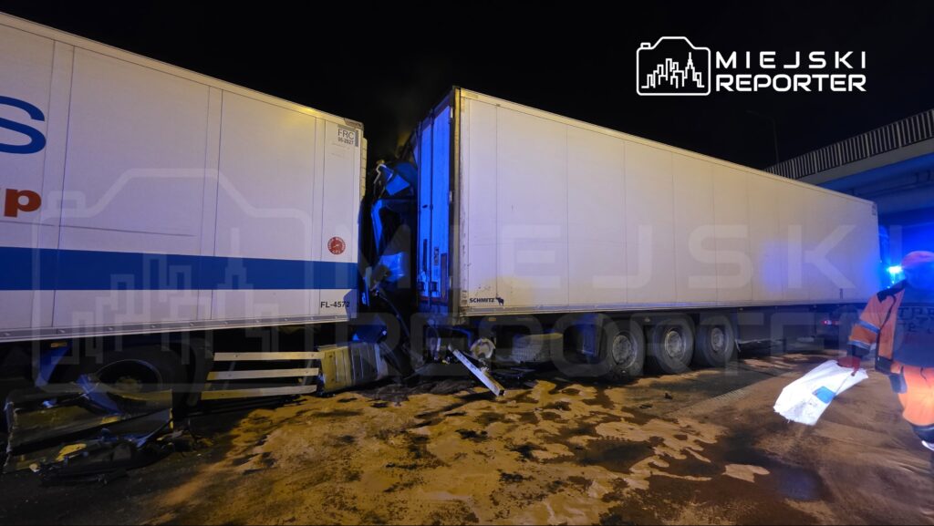 Zderzenie dwóch ciężarówek na drodze, z uszkodzeniami naczep i leżącymi na ziemi elementami. W tle widać pracownika w kamizelce odblaskowej.