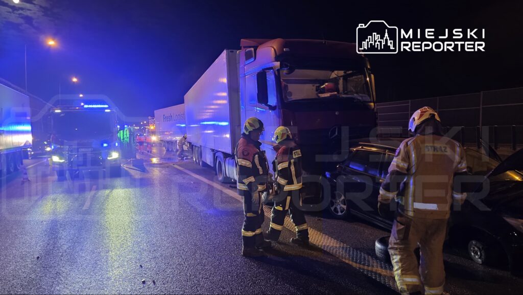 Grupa strażaków w odblaskowych strojach ocenia skutki wypadku drogowego na nocnej drodze, obok ciężarówek.
