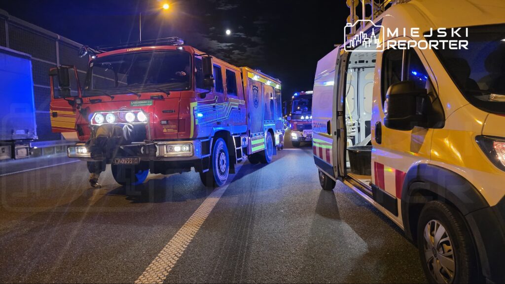 Czerwony wóz strażacki z włączonymi sygnalizatorami stoi na drodze nocą, obok karetka z otwartymi drzwiami.