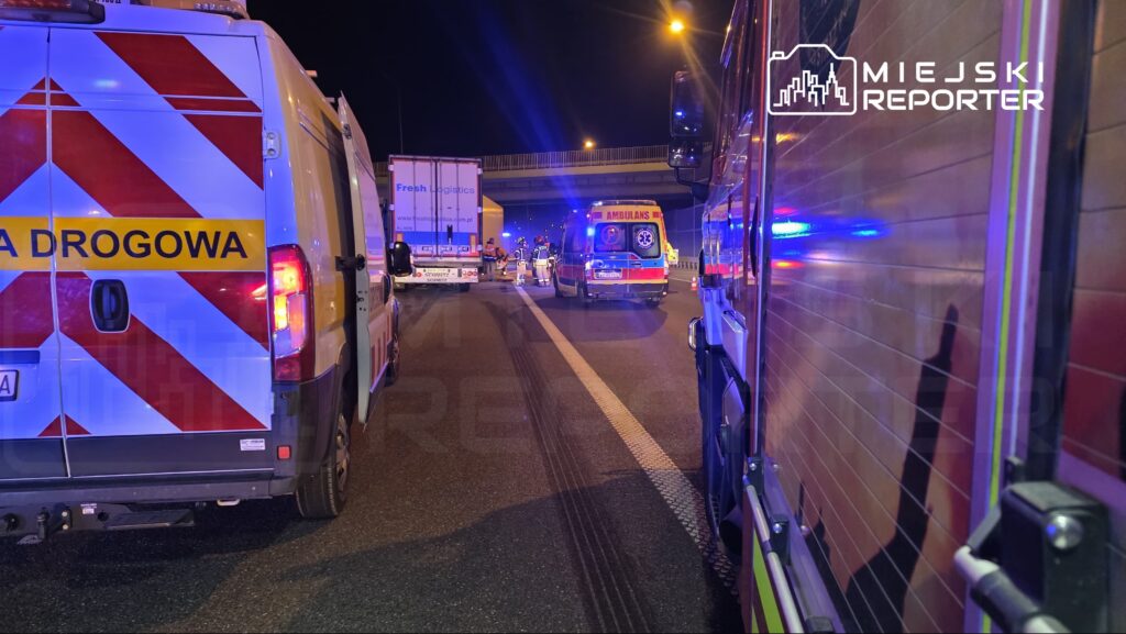 Karetka pogotowia oraz pojazdy służb ratunkowych zatrzymane na drodze, w nocy, przy oświetleniu ulicznym.