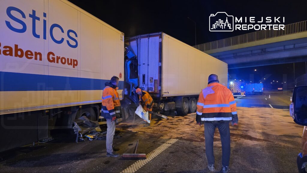 Czterej pracownicy w pomarańczowych kurtkach zabezpieczają miejsce wypadku drogowego z udziałem ciężarówki.