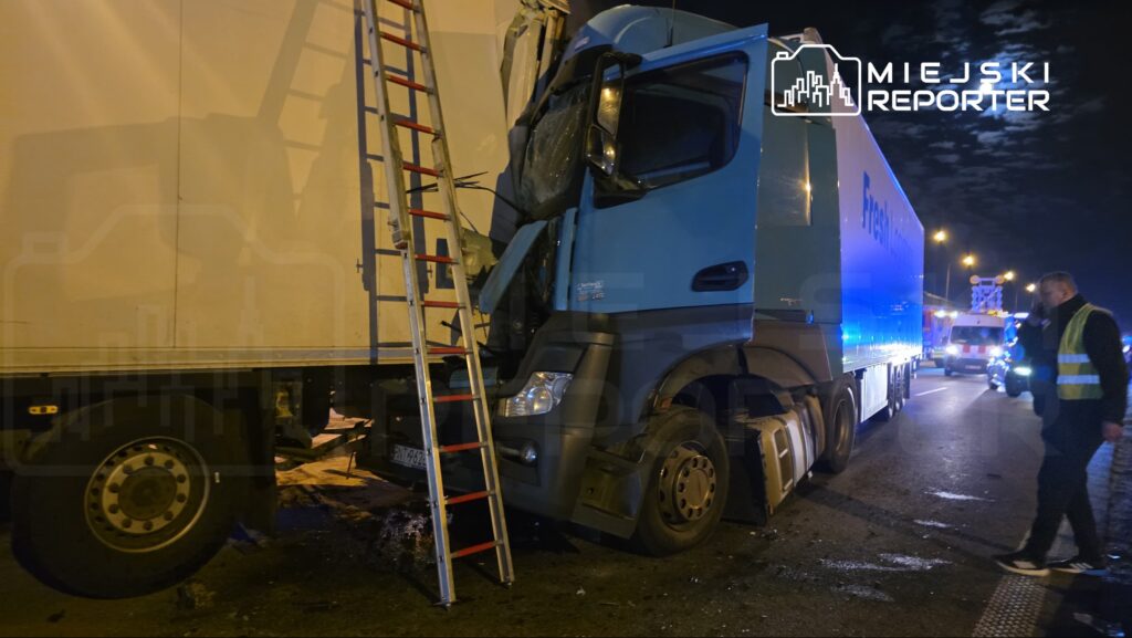 Czarny ciężarowy samochód z uszkodzoną kabiną stoi przy drodze, obok niego drabina oraz mężczyzna w kamizelce odblaskowej.