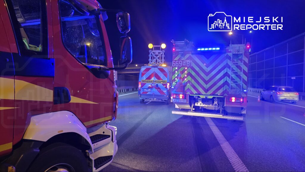 Na drodze nocą stoją dwa pojazdy służbowe: strażacki i drogowy, z włączonymi sygnałami świetlnymi.