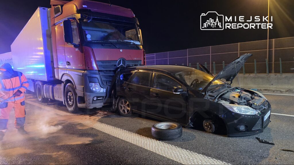 Ciężarówka z czerwoną kabiną stoi obok uszkodzonego czarnego samochodu osobowego z otwartą maską na drodze.