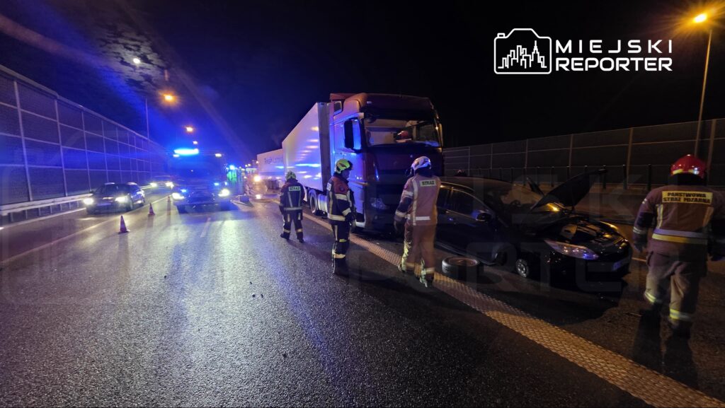 Grupa strażaków w mundurach bada uszkodzone samochody na drodze w nocy, obok stoi wóz strażacki z włączonymi sygnałami.