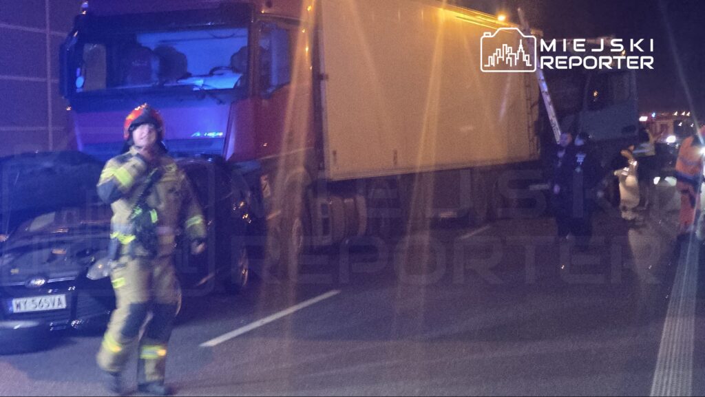 Strażak w mundurze z hełmem, wskazujący na ciężarówkę na drodze, otoczoną przez inne pojazdy ratunkowe.