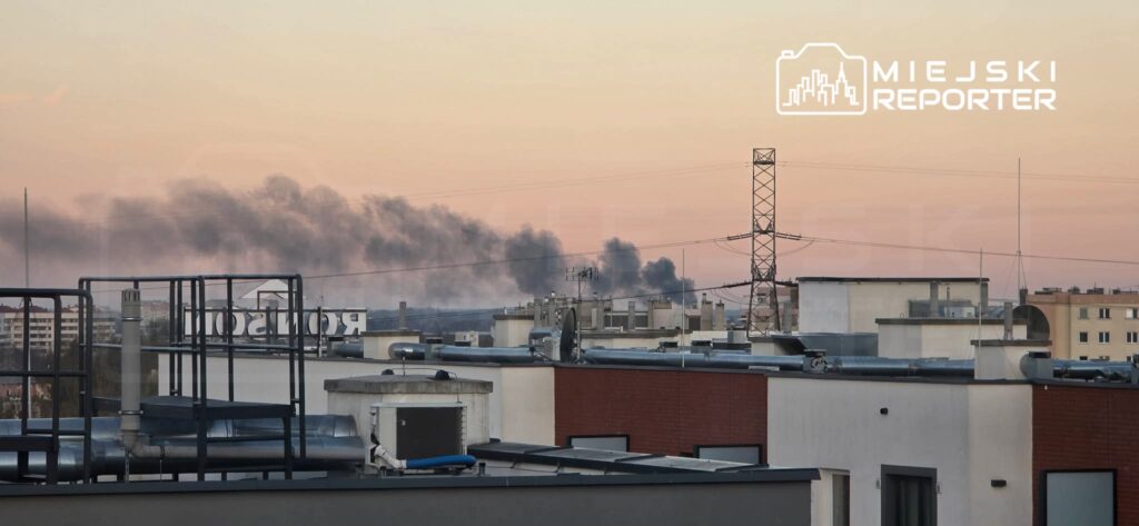 Na tle zachodzącego słońca, z dachów budynków unosi się czarny dym, a w oddali widoczna jest wieża energetyczna.
