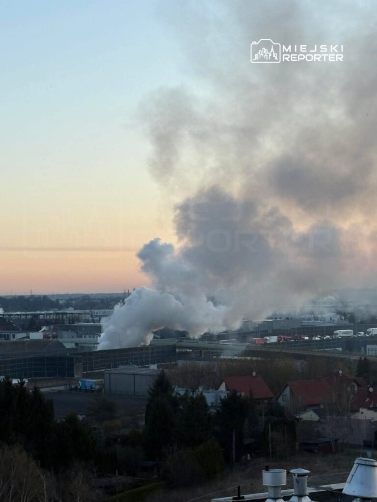Gęsty dym unosi się z kominów przemysłowych nad miastem o zmierzchu, w tle widać budynki i samochody na drodze.
