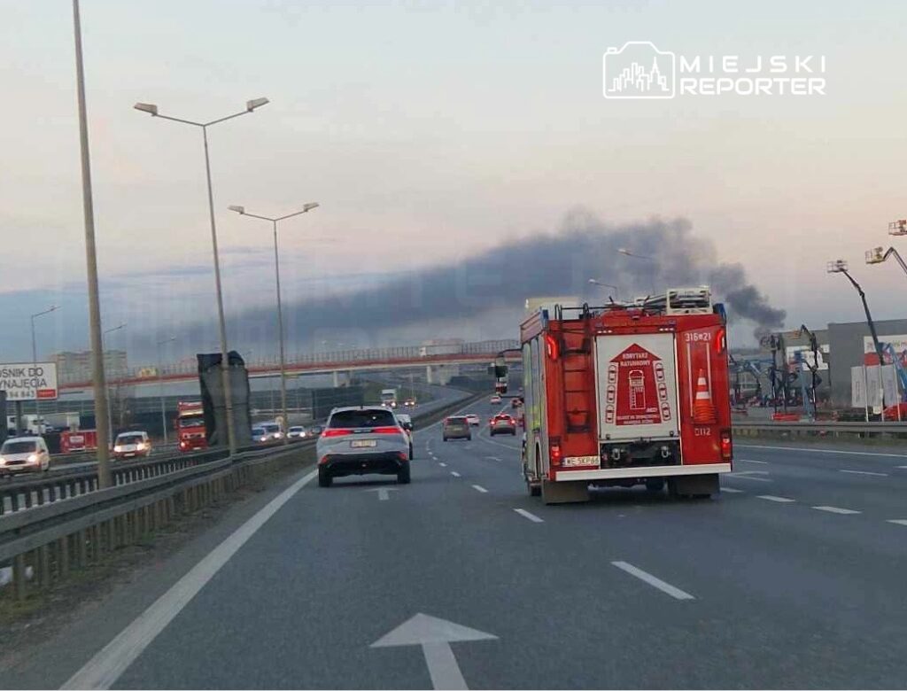 Czerwony wóz strażacki porusza się autostradą, w tle widoczny czarny dym unoszący się nad miastem.