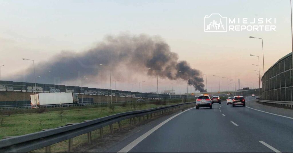 Gęsty, czarny dym unoszący się nad autostradą, w otoczeniu pojazdów osobowych i ciężarowych.