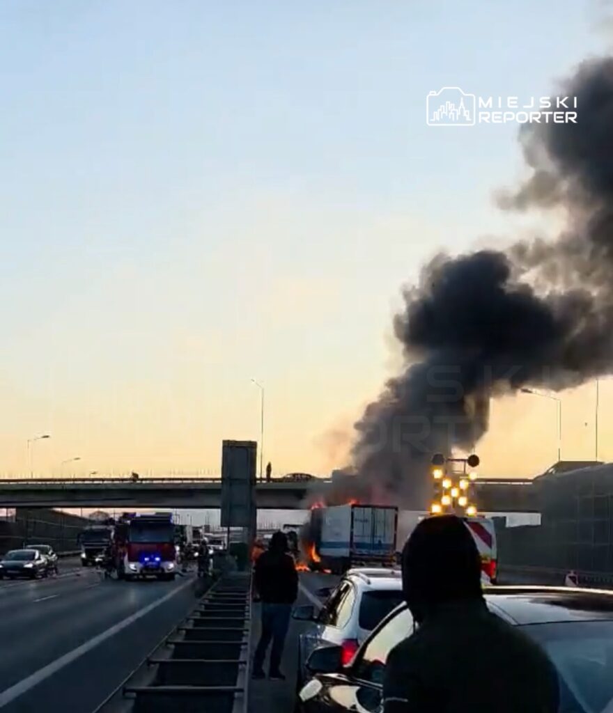 Zapalony samochód ciężarowy na autostradzie, z unoszącym się czarnym dymem, obok pojazdy i straż pożarna.