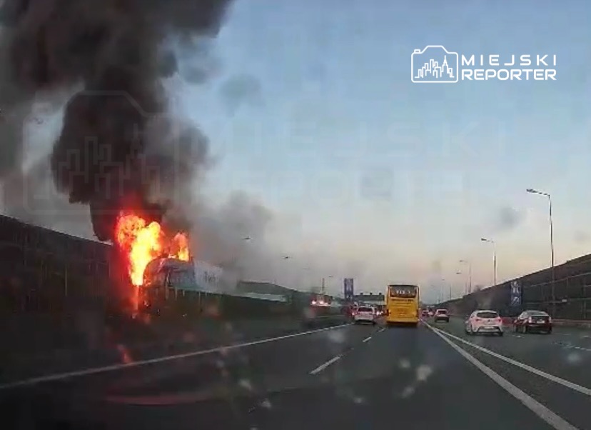 Pojazd płonący na autostradzie, z dużym kłębem czarnego dymu unoszącym się w powietrze. Ruch samochodowy w tle.