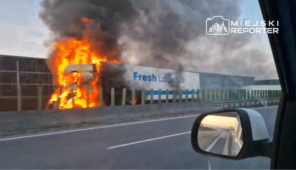 Tir płonący na poboczu drogi, wydobywający dym i płomienie, obok nowoczesny budynek magazynowy.
