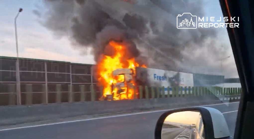 Ciężarówka płonąca przy drodze, otoczona gęstym dymem, z dużymi płomieniami wydobywającymi się z jej wnętrza.