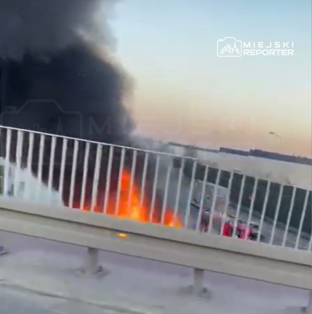 Pojazd płonący na poboczu drogi, z dużym słupem czarnego dymu unoszącym się w powietrze.