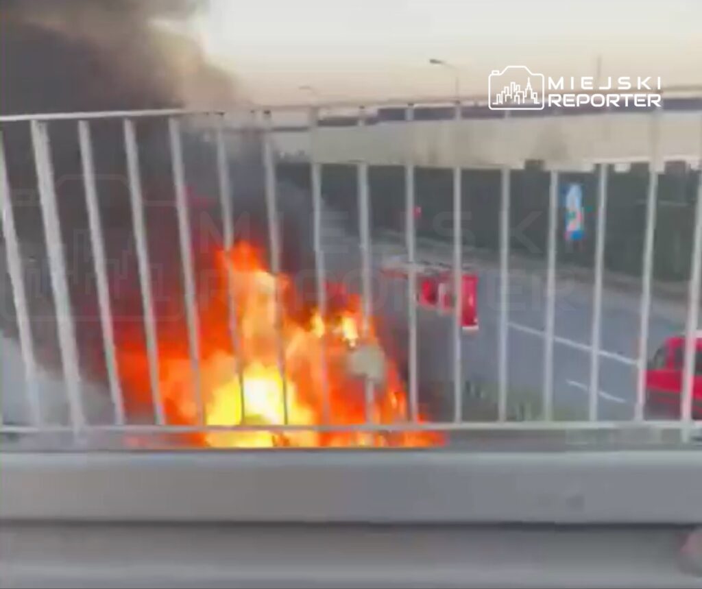 Pojazd płonący na poboczu drogi, otoczony gęstym dymem, w tle widoczne inne samochody.