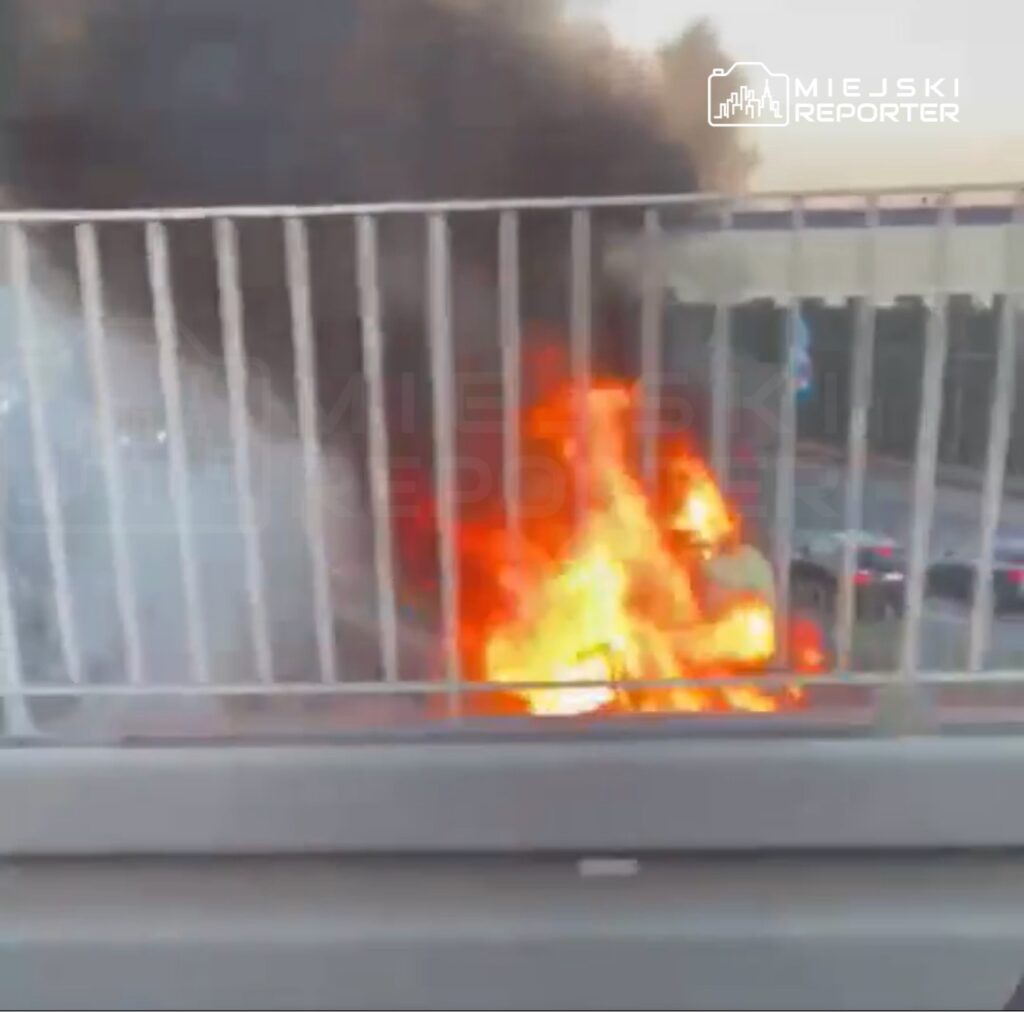 Ogień i dym wydobywają się z płonącego obiektu za barierką na moście, w tle widoczne samochody.