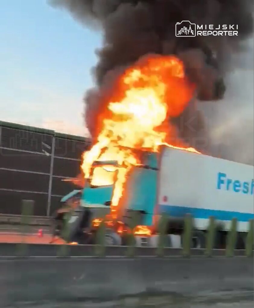 Cysterna ciężarówki płonie na drodze, unosząc się czarny dym w tle.
