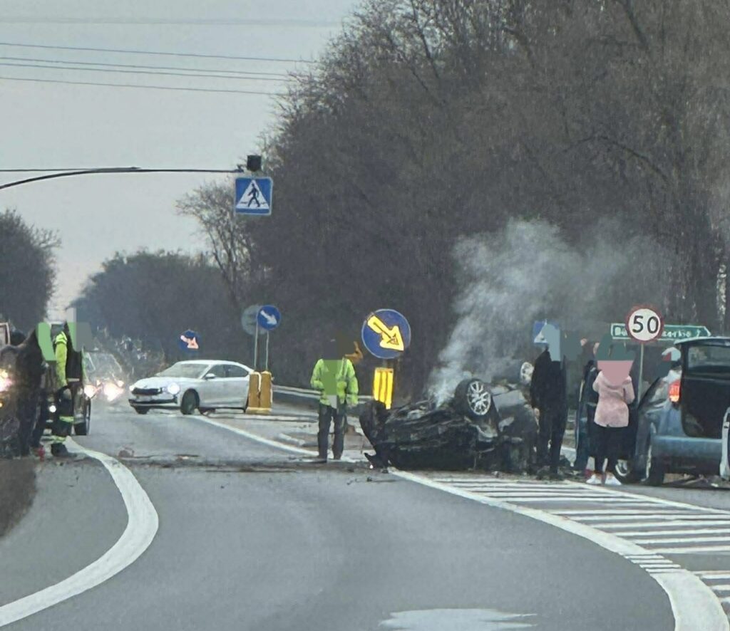 Na drodze leży przewrócony samochód, z którego wydobywa się dym; obok stoją strażacy i gapie.