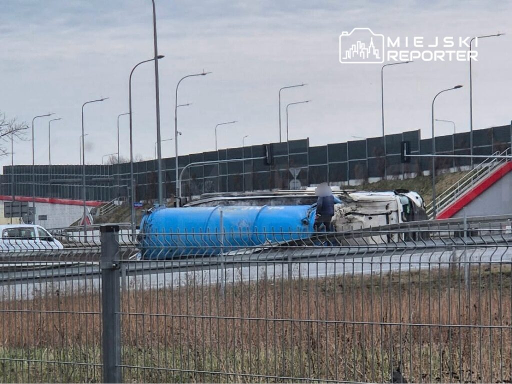 Przewrócony niebieski pojazd ciężarowy na jezdni, obok stoi mężczyzna w ciemnej odzieży, w tle nowoczesny budynek.
