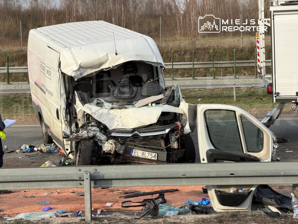 Zniszczony furgon po wypadku drogowym, obok którego stoi policjant z notatnikiem; na jezdni leżą szczątki i odpadki.